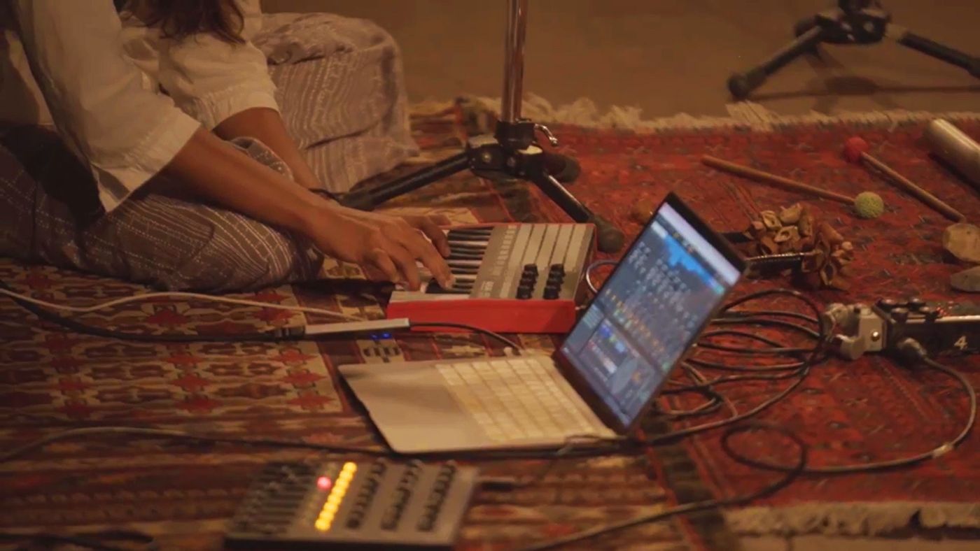 Hands on a MIDI keyboard beside a laptop running Ableton, on a rug.