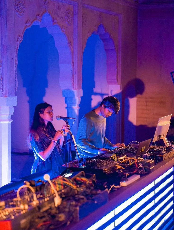 Sahitya singing and Harsh at a modular synth rig under violet light, blue-and-white striped stage front.