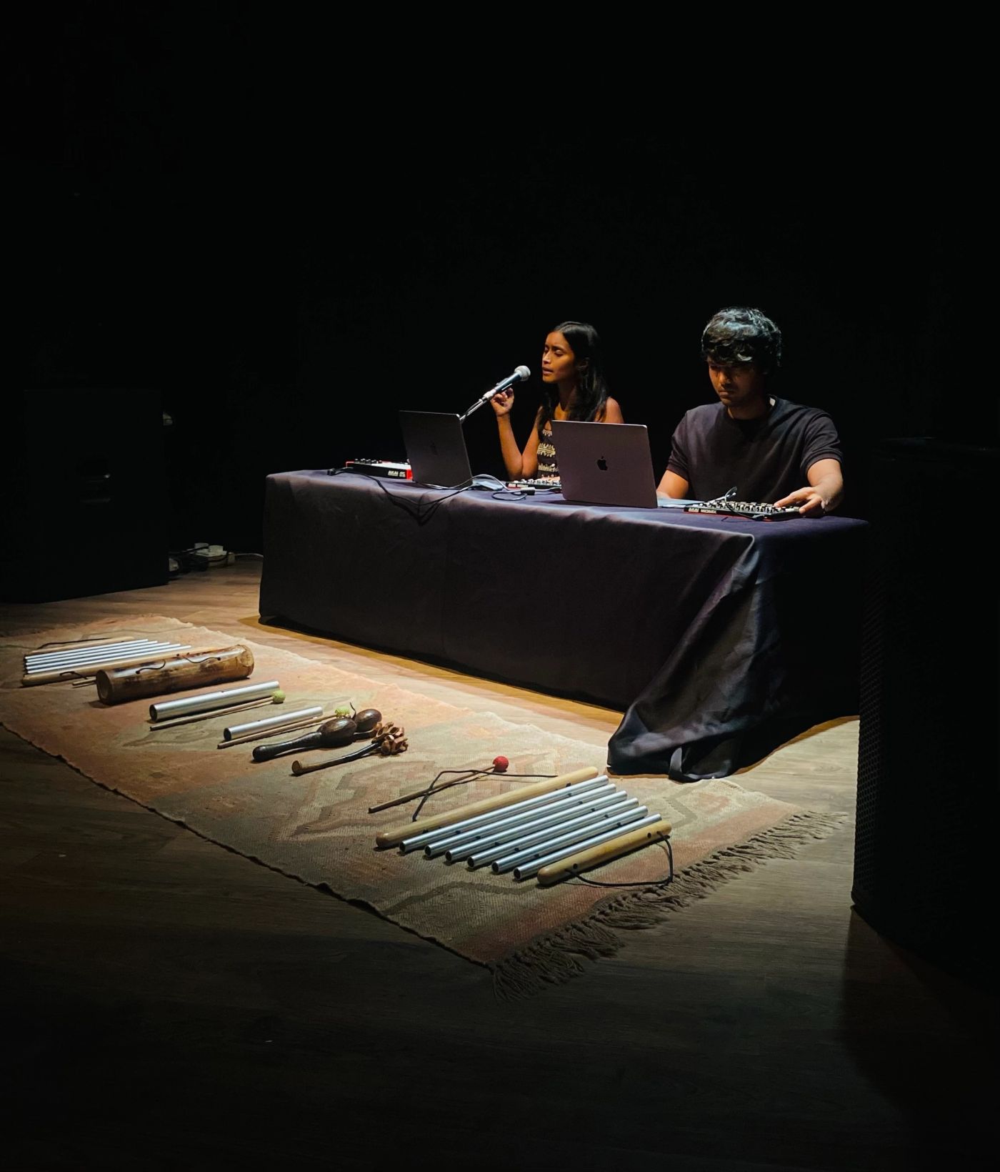 Sahitya and Harsh on a dim stage, chimes and percussion laid out on a rug.