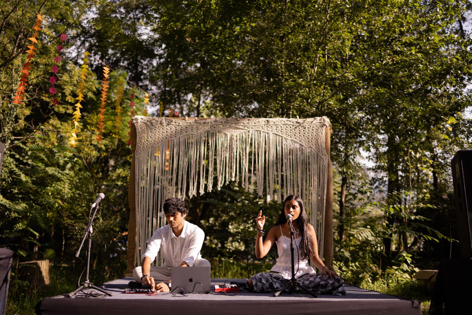 Harsh at a laptop and controller and Sahitya singing, seated on a small outdoor stage under a macramé canopy among trees at GAIA Festival.